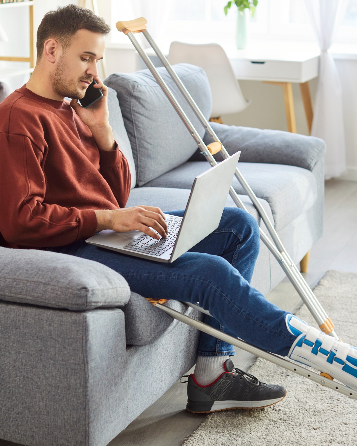 A man with a leg cast sits on a sofa, using a laptop and talking on the phone. Crutches lean beside him. The room is bright and modern.
